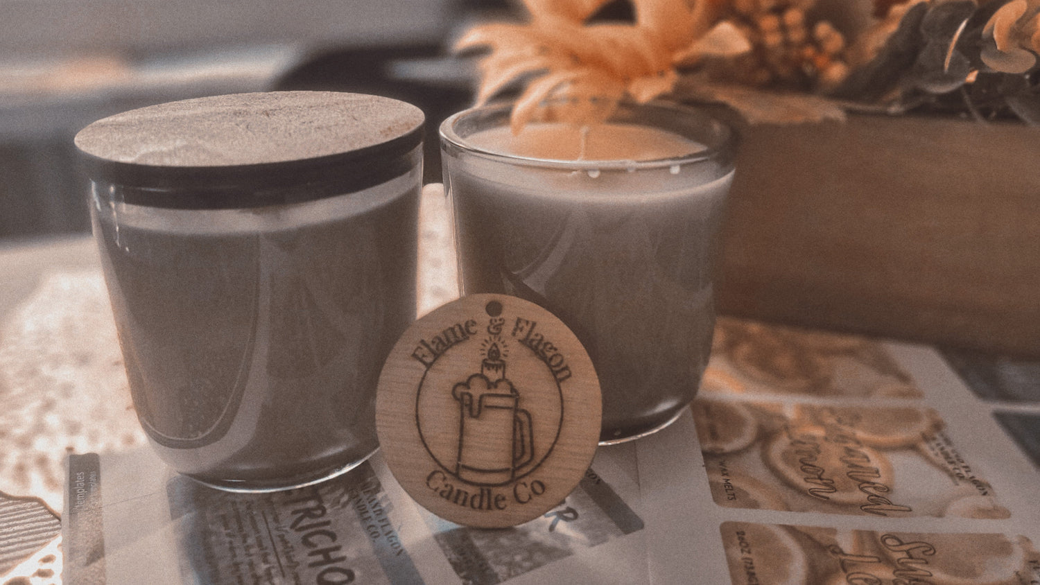 Two candles in glass containers with a wooden coaster featuring 'Flame & Flagon Candle Co' on a pile of candle labels.